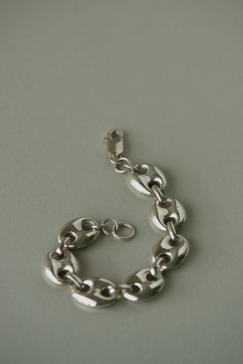Silver puffed marine link bracelet arranged in a gentle curve against a sage green background. The highly polished oval links catch light along their rounded edges, creating a rhythmic pattern of reflections that emphasizes the piece's substantial yet fluid form. by Sydney SG
