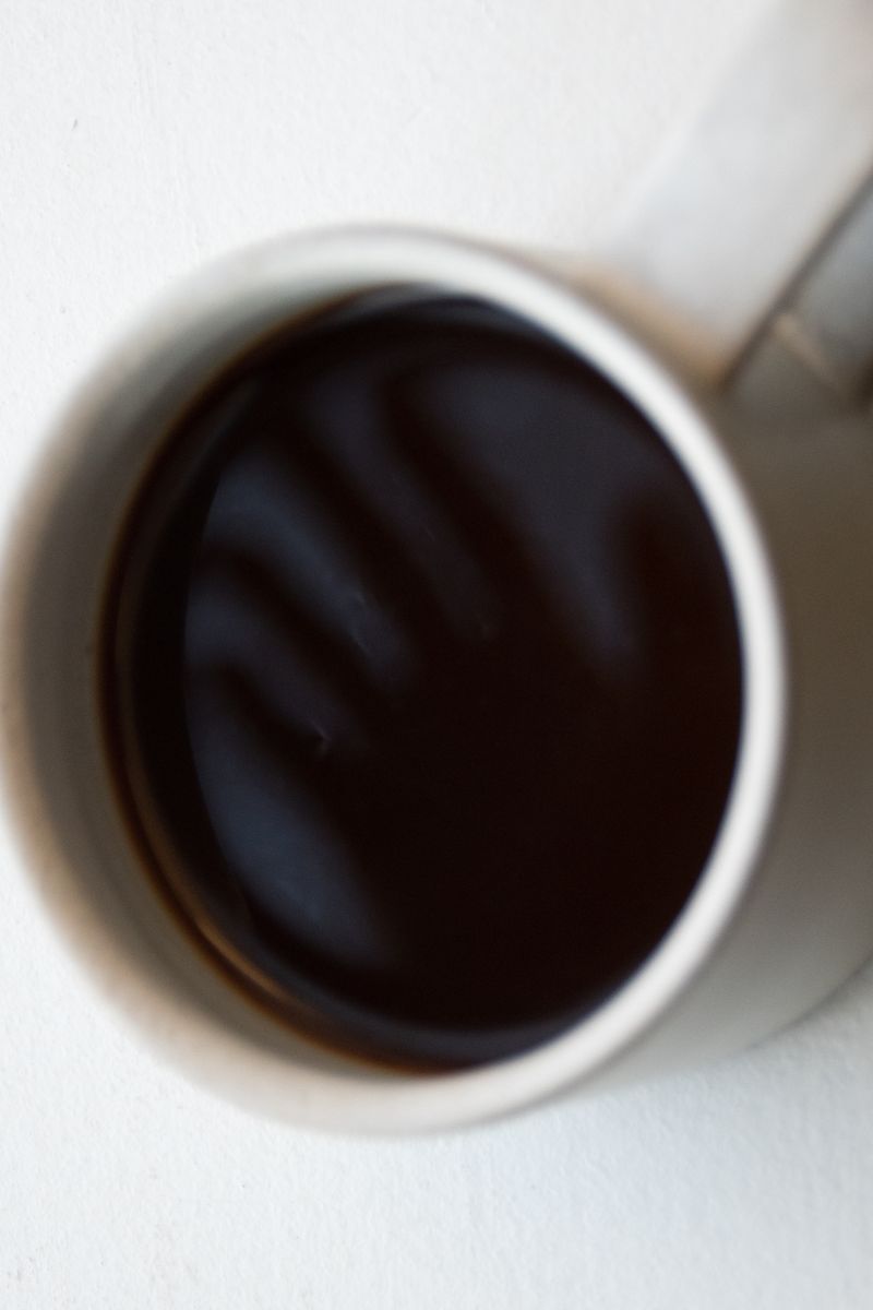 Overhead view of black coffee in a white ceramic cup reflecting the silhouette of an opened hand. Photographed with intentional blur. The stark contrast and circular form create a hypnotic, contemplative focal point. by Sydney SG