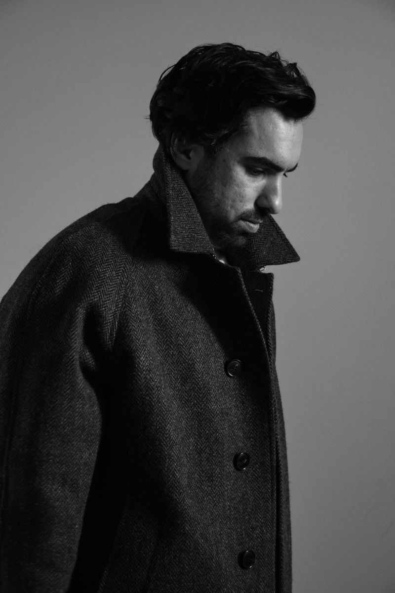 Monochromatic portrait of a man in profile, head slightly bowed, wearing a textured herringbone coat with upturned collar. The dramatic shadows and somber expression create a pensive, cinematic atmosphere. by Sydney SG