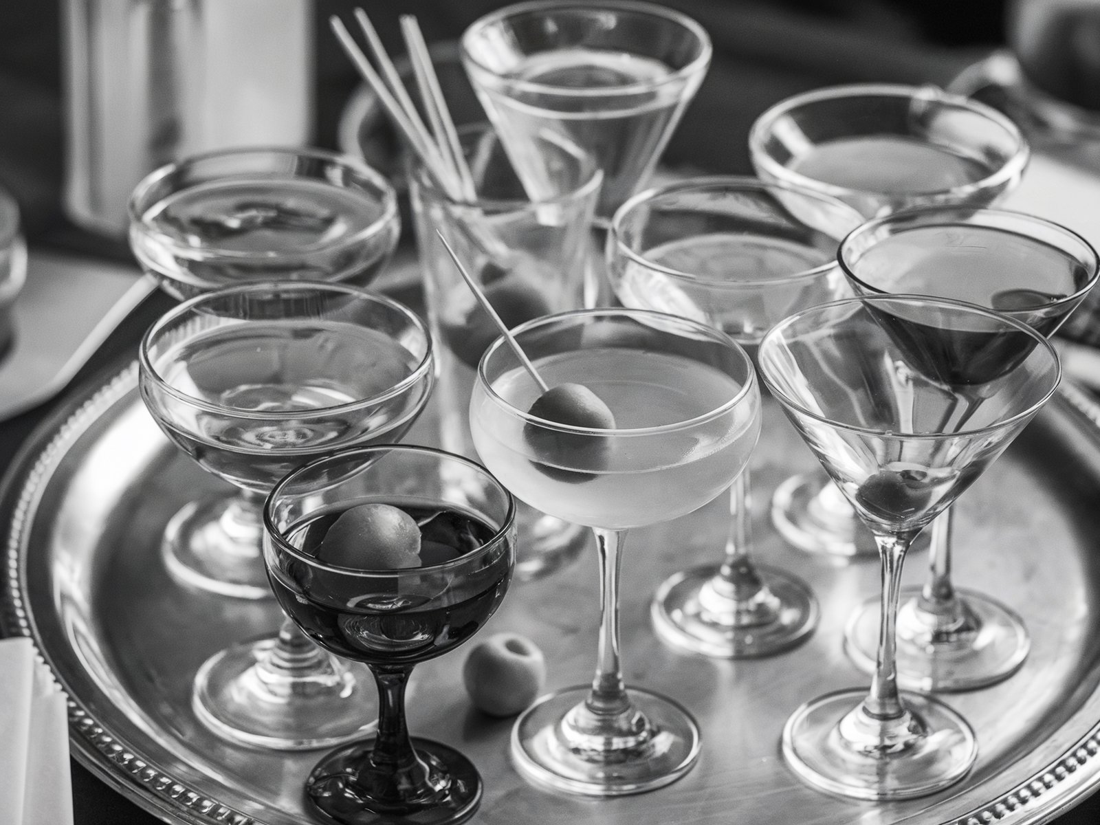 Black and white photo of a tray filled with cocktail glasses, toothpicks, and olives. The arrangement exudes a sophisticated, classic cocktail hour vibe. Image generated with AI. by Sydney SG