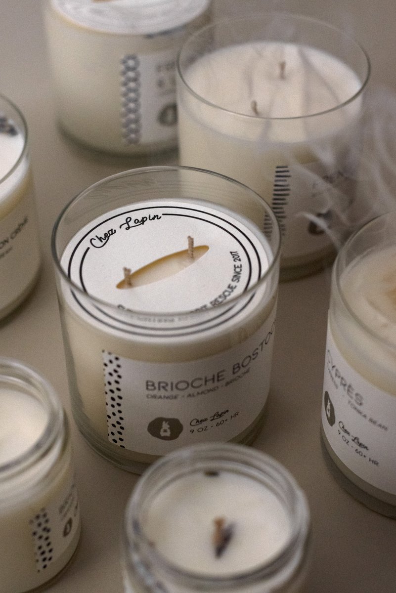 Group of scented candles in glass jars, some of which have visible labels and delicate smoke wisps rising from recently extinguished wicks. by Sydney SG