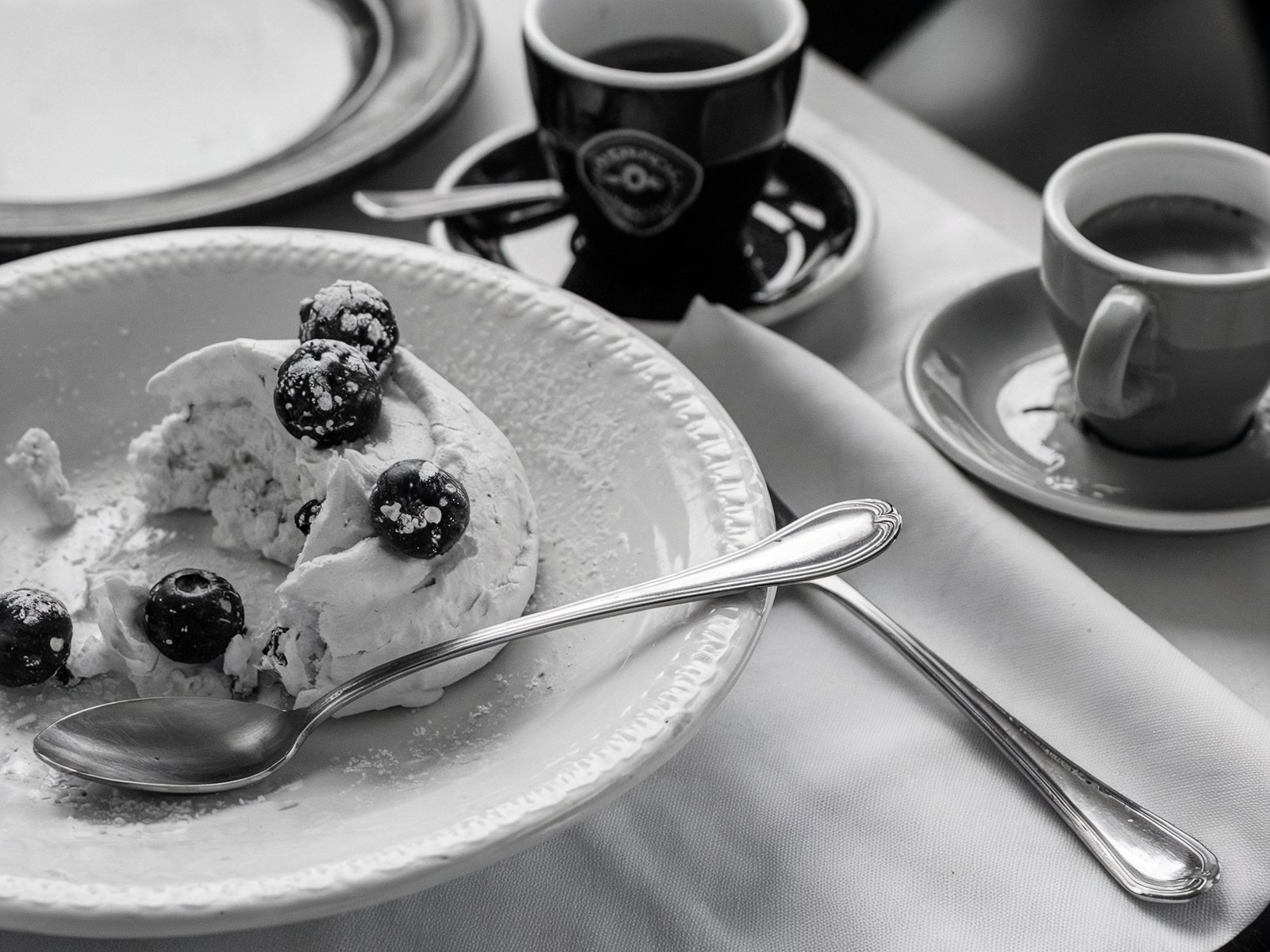 Monochromatic image of a partially eaten pavlova with berries on a white plate, accompanied by two espresso cups. The scene captures a quiet moment of indulgence. Image generated with AI. by Sydney SG