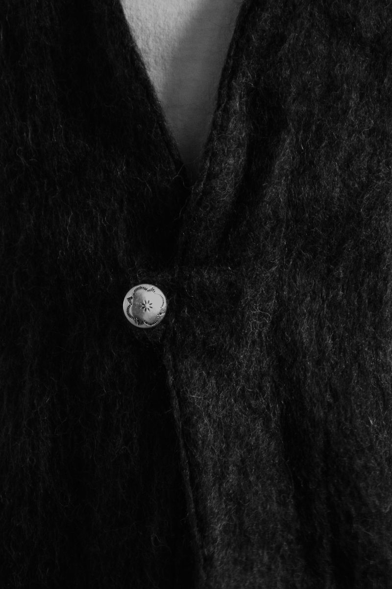 Closeup of a dark wool garment with a delicate silver button. The monochromatic palette emphasizes the textural qualities of the fabric and the crafted detail of the metallic closure. by Sydney SG
