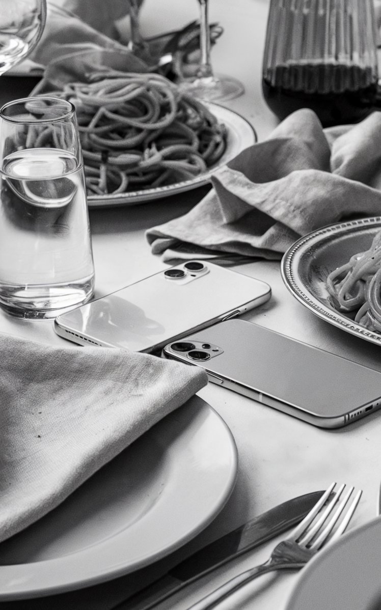 Black and white photo of an table loaded with plates of pasta and glasses of wine and water, and two abandoned cell phones laying face down on the tablecloth. A close up of a dinner table surrounded by diners in conversation. Image generated with AI. by Sydney SG