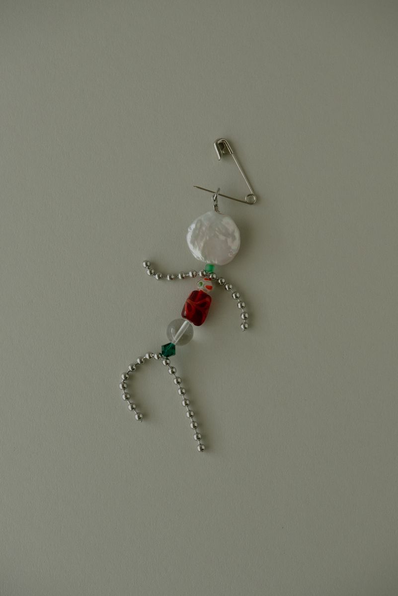 Whimsical silver figurine arranged from ball chain and beads, resembling a stick figure with a lustrous pearl disc head, vibrant red cube torso, and clear crystal accent. The safety pin attachment adds playful functionality to this handcrafted character pendant. by Sydney SG