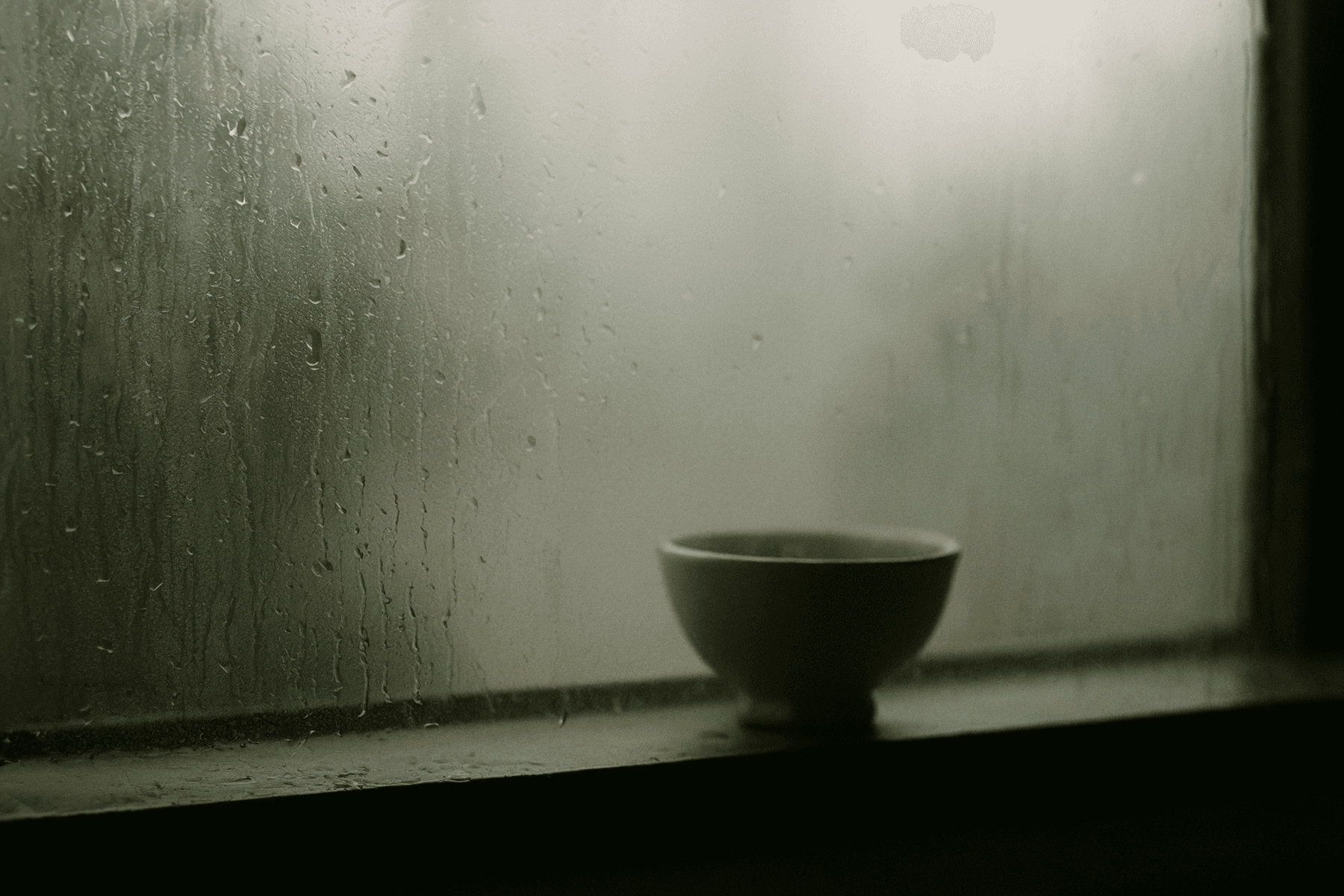 A dark ceramic bowl sits alone on a windowsill with rain droplets covering the glass. Soft gray light filters through the wet window creating a contemplative mood. (AI-generated image) by Sydney SG