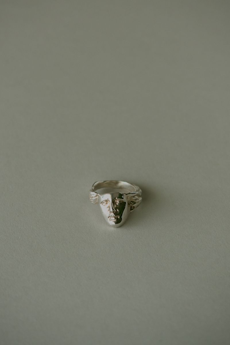 Sterling silver face ring centered on a pale sage background. The sculptural portrait shows half in polished brightness, half in shadow, creating a dramatic duality while the textured band wraps organically around the form like embracing hands. by Sydney SG