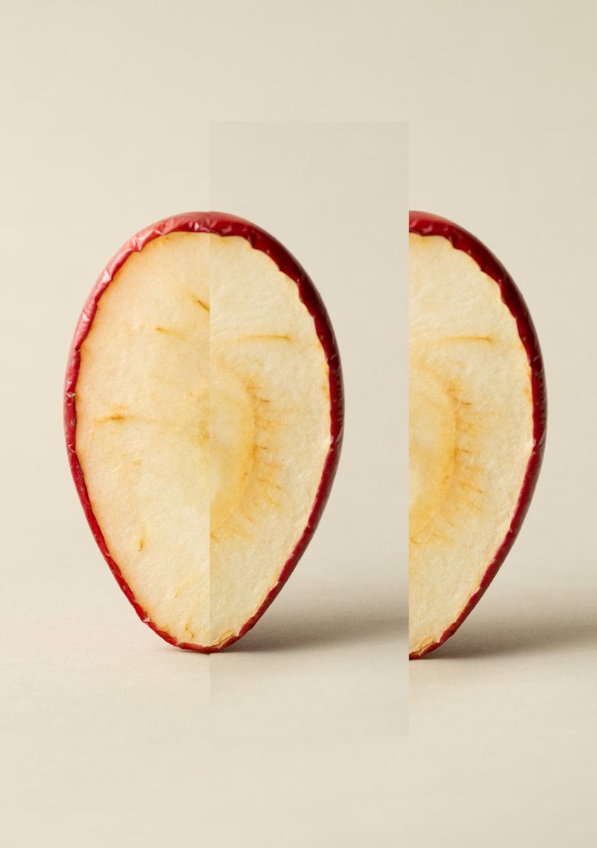 An apple slice with a visible split down the center, revealing a creamy white interior with a thin red skin edge. The background is a neutral beige. by Sydney SG