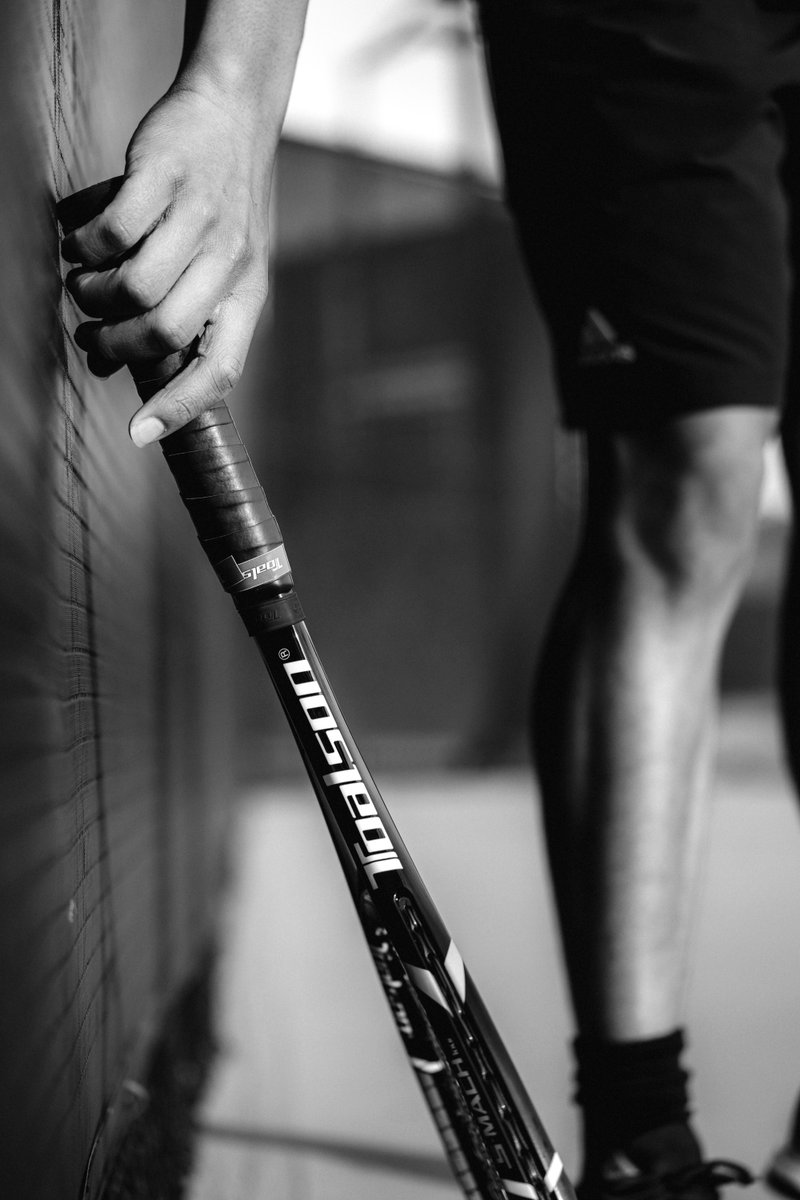 Person picking up a tennis racket that's leaning against a court fence, shown in black and white with a focus on the hand and racket. by Sydney SG