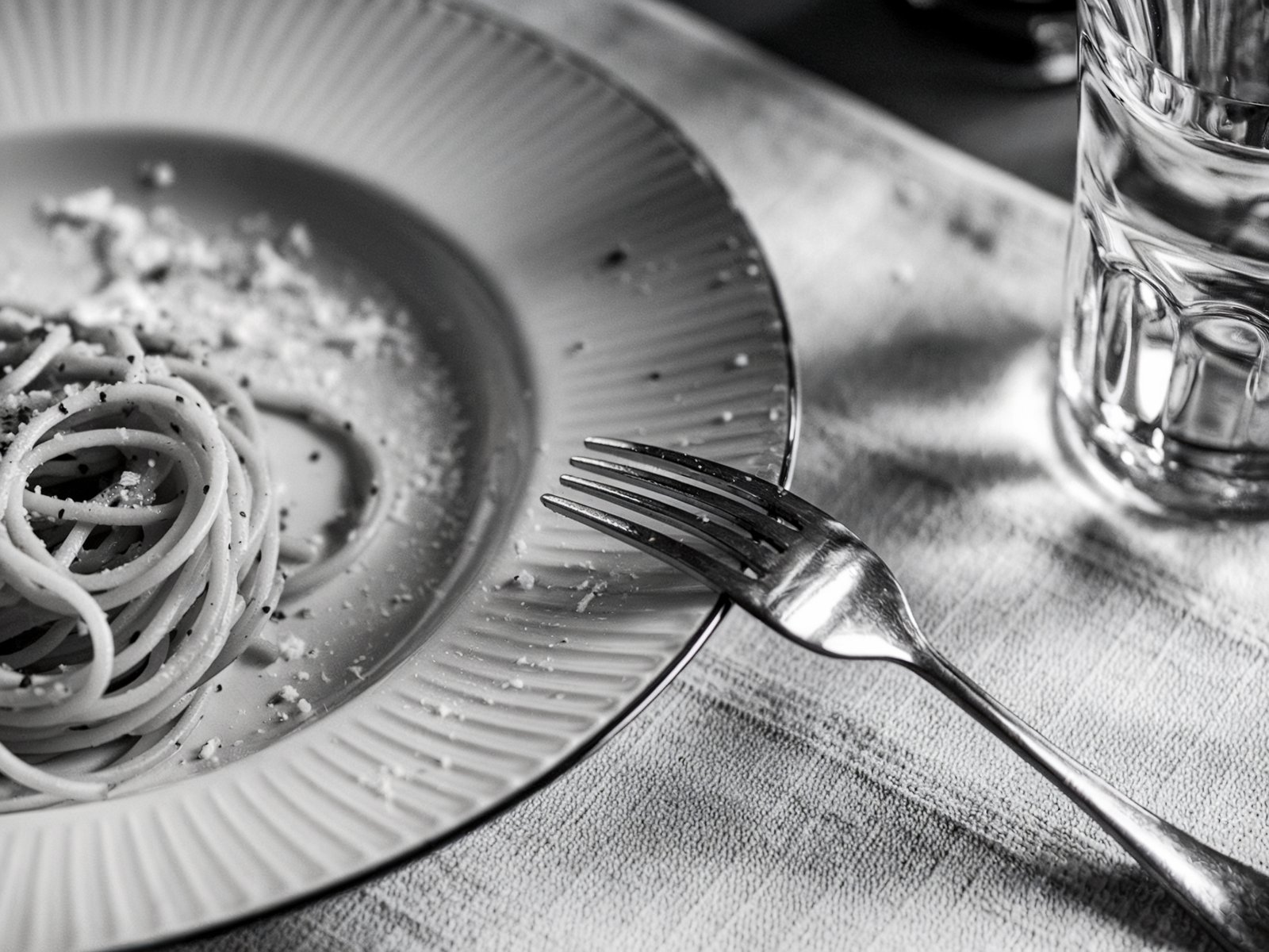 Monochrome image of a plate with spaghetti and scattered cheese crumbs, accompanied by a fork and a glass of water. A minimalist table setting with a casual dining atmosphere. Image generated with AI. by Sydney SG