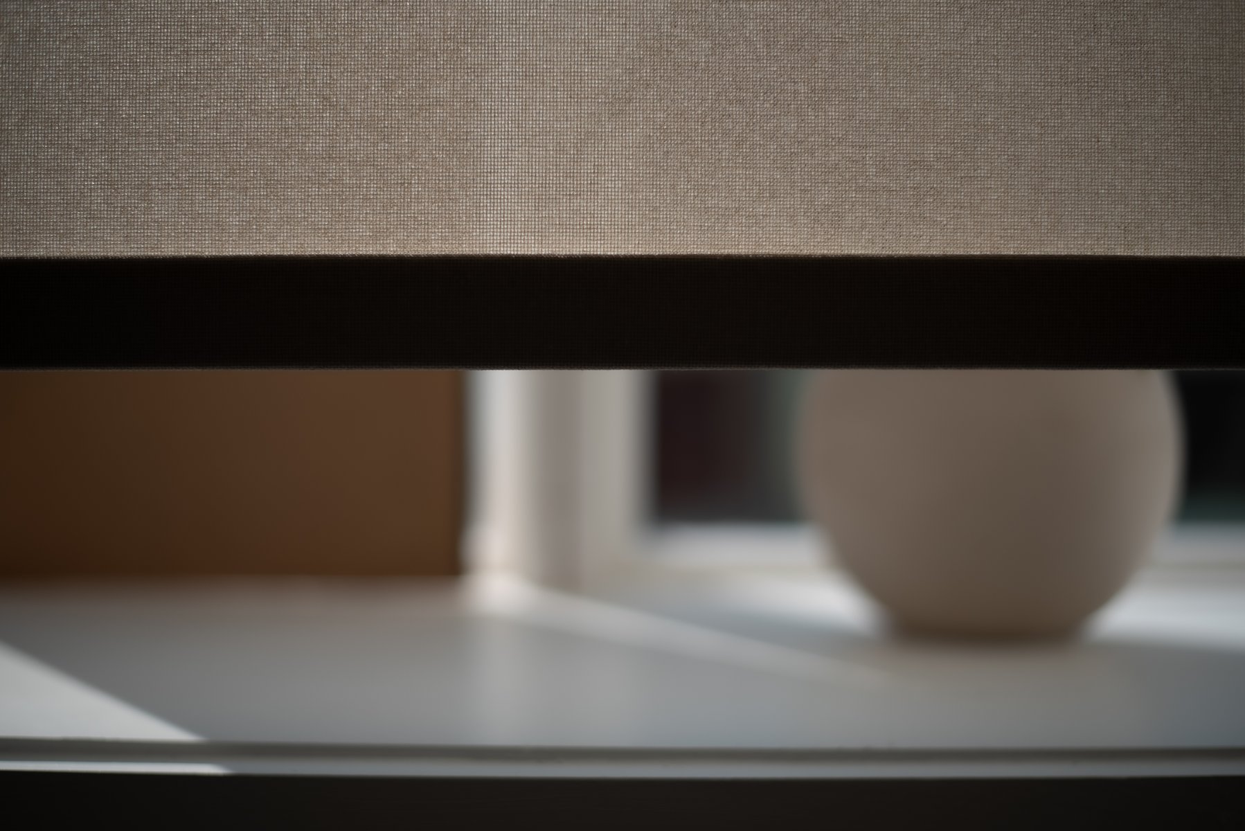 Close-up of a partially open window shade casting a shadow, with a blurred round object in the background, suggesting depth and light play. by Sydney SG