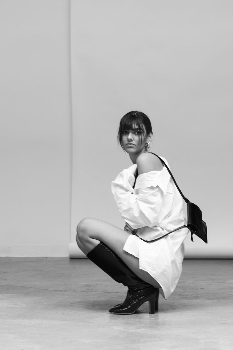 Monochromatic portrait of a woman sitting on her heels while wearing an oversized white shirt and knee-high black boots, with a small black handbag hanging over her shoulder by Sydney SG