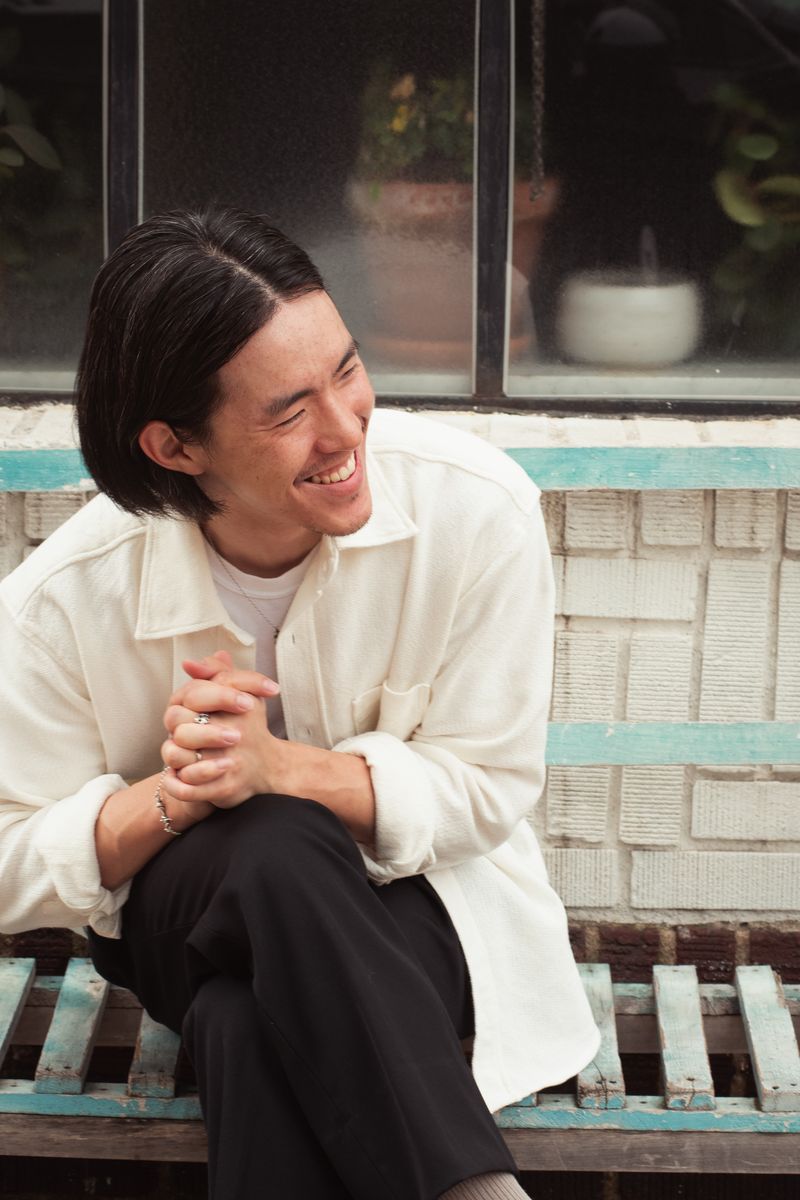 A person in a white shirt laughing while sitting on weathered wooden steps with turquoise paint. The candid moment captures genuine joy against the rustic, colorful architectural detail. by Sydney SG