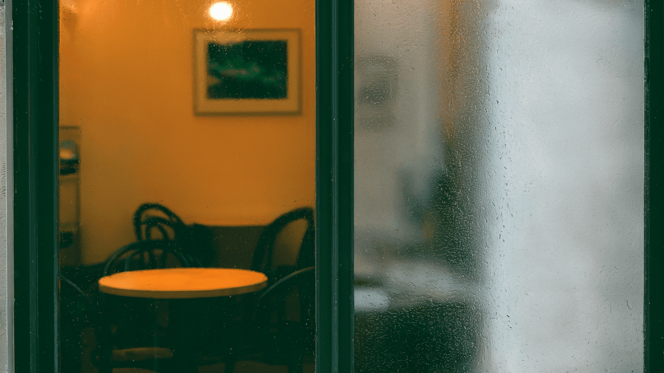 Looking through a rain-covered glass door toward a warm, amber-lit café interior with green trim. The glass distorts the view of the round table and chairs inside. (AI-generated image) by Sydney SG