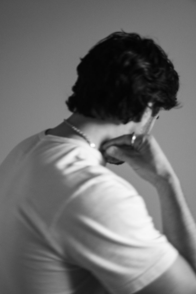 Soft-focus portrait of a man seen from behind, wearing a white t-shirt with delicate silver chain visible. The monochromatic treatment and gentle blur create a sense of vulnerable intimacy. by Sydney SG