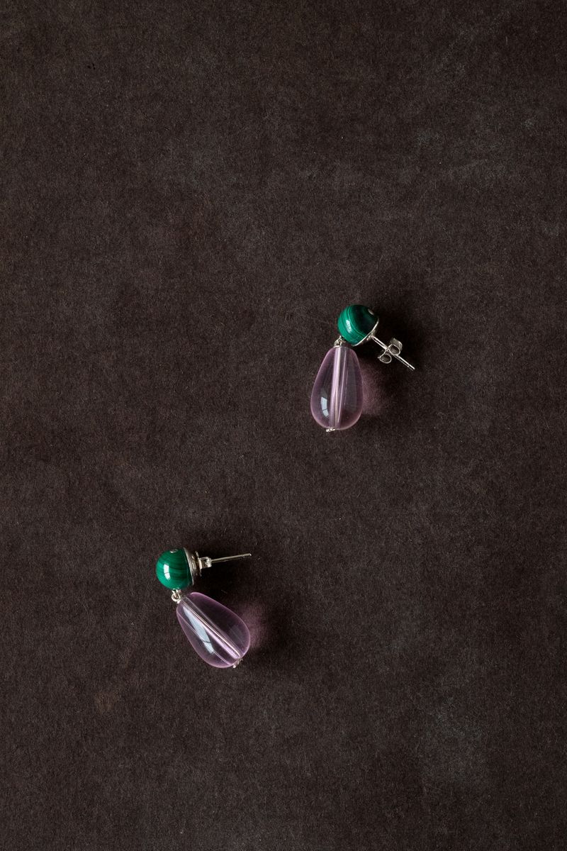 A pair of earrings photographed against a dark charcoal background, showing green cabochon studs with pink translucent drops. The dramatic lighting emphasizes the gemstones' natural translucency and rich color saturation. by Sydney SG