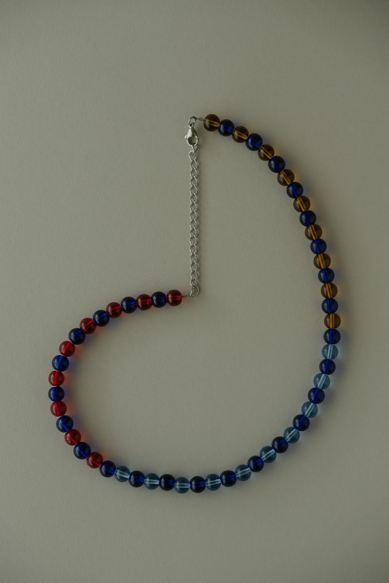 Beaded necklace featuring alternating cobalt blue, amber, and red glass beads with silver chain extension. The rhythmic pattern of translucent spheres creates a playful color story as light passes through each bead, revealing their varied internal luminosity. by Sydney SG