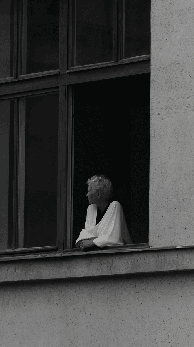 Black and white photograph of a person in a white shirt sitting at an open window, looking outward. The architectural framing and contemplative pose create a sense of quiet observation from within. (AI-generated image) by Sydney SG